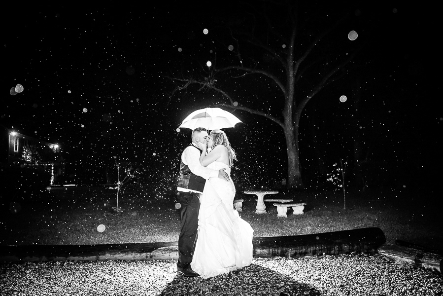 A night shot of a bride and groom kissing under an umbrella as it rains