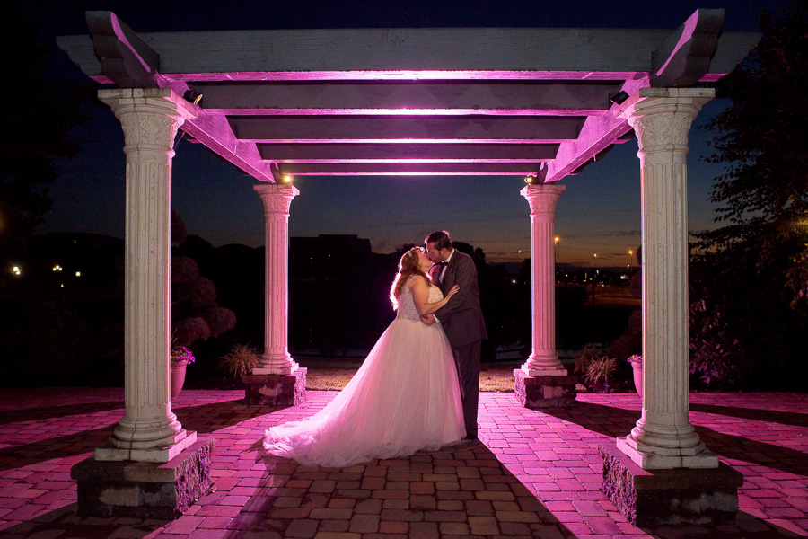 Night photo of bride and groom kissing under the pergola as purple light is popped behind them