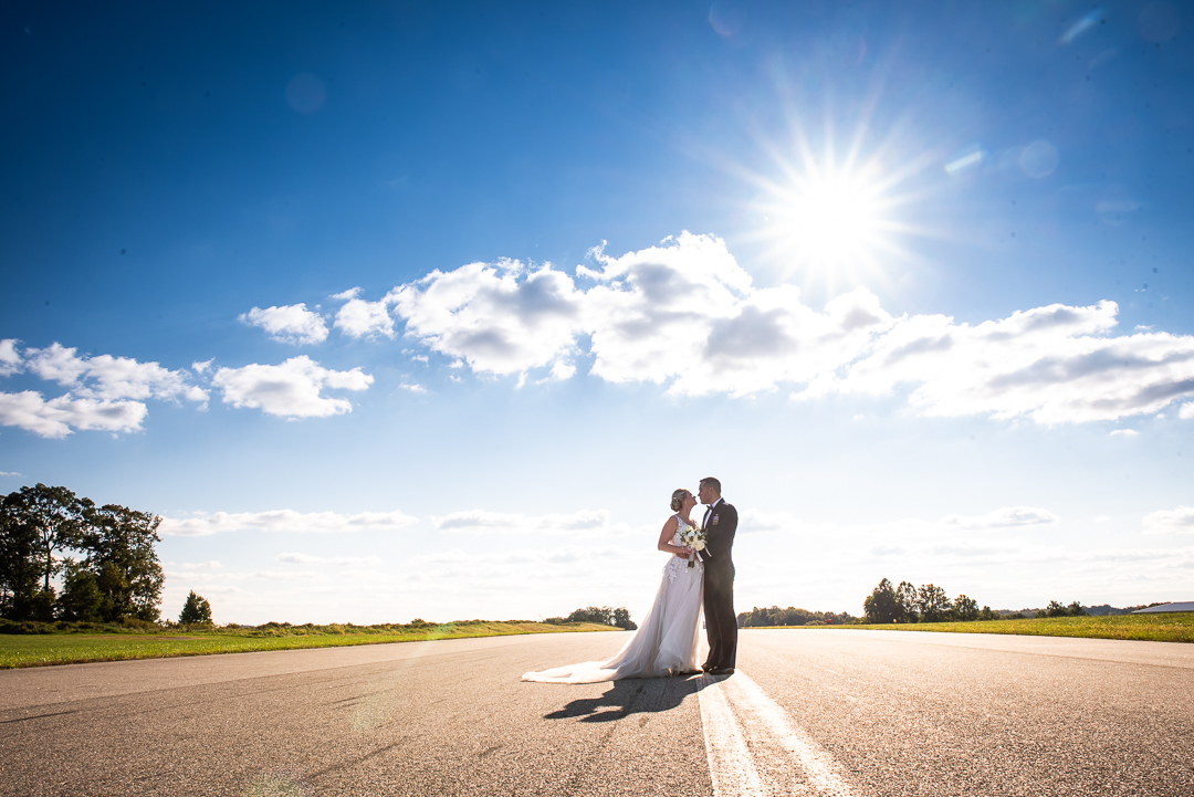 bride and groom almost kissing on runway with sun and clouds in background after airport wedding ceremony