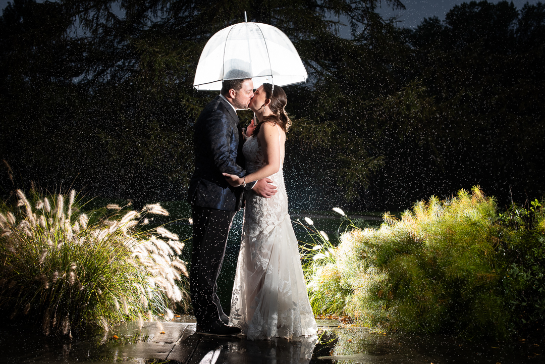 Mid-october wedding photo of bride and groom under umbrella at night with light behind them and rain falling
