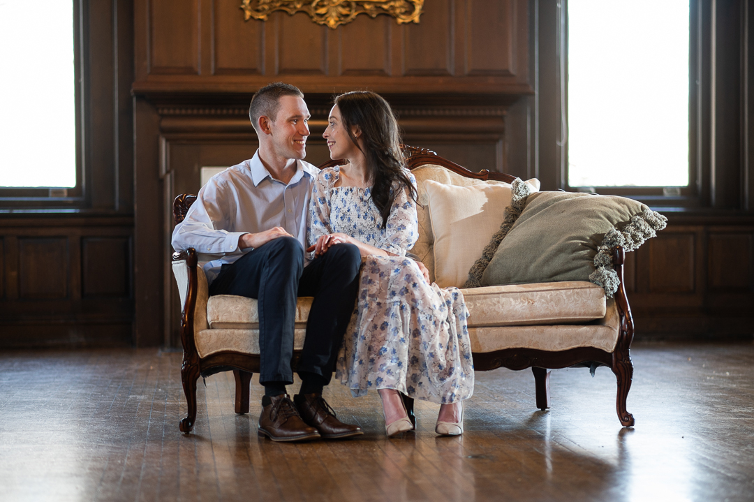 indoor engagement session photo of couple sitting on victorian couch smiling at each other